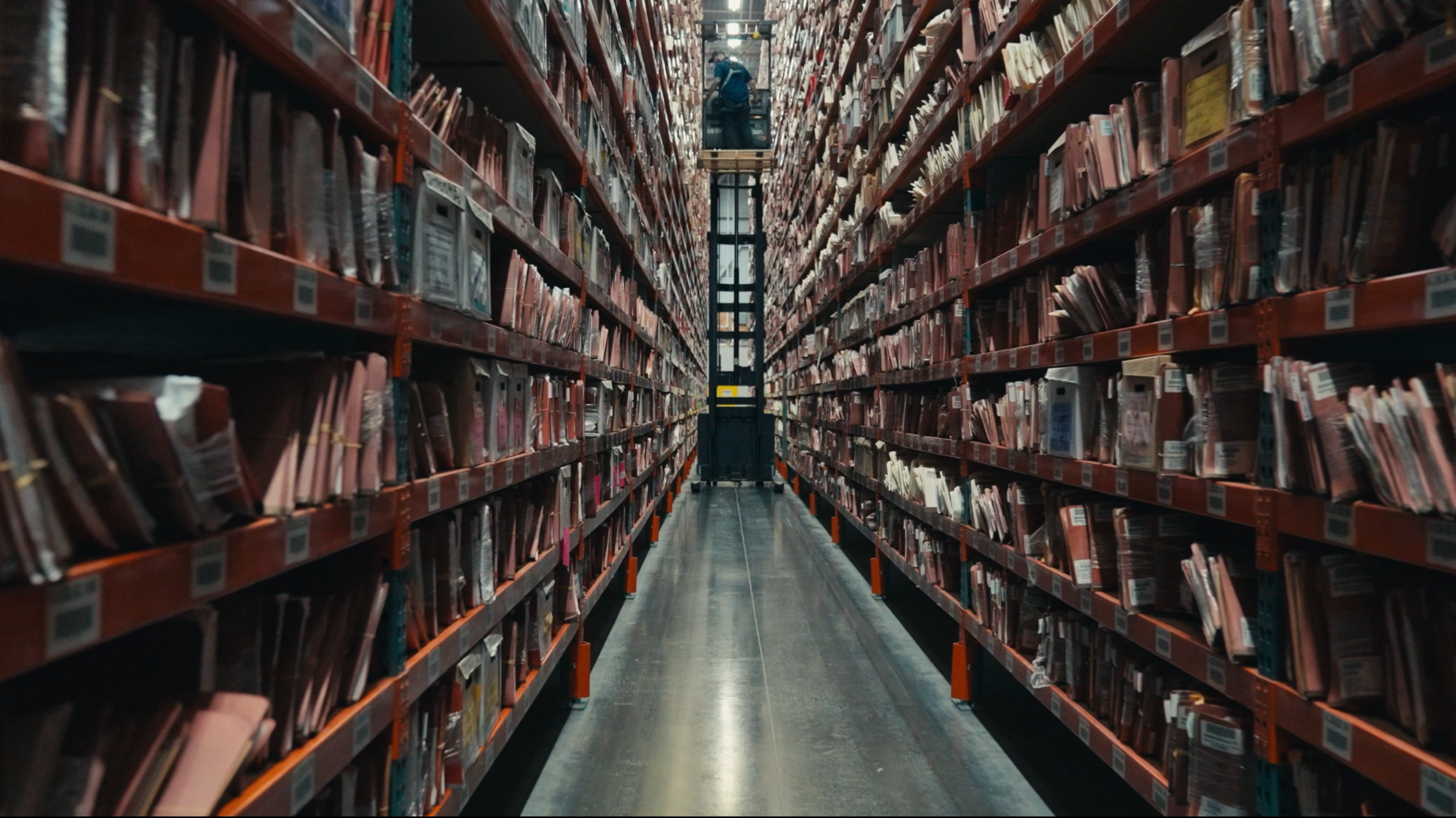An image of the archive stacks, a long hallways with shelves going up to the ceiling and an archivist using machinery to move down the hallway at the top of the stacks.