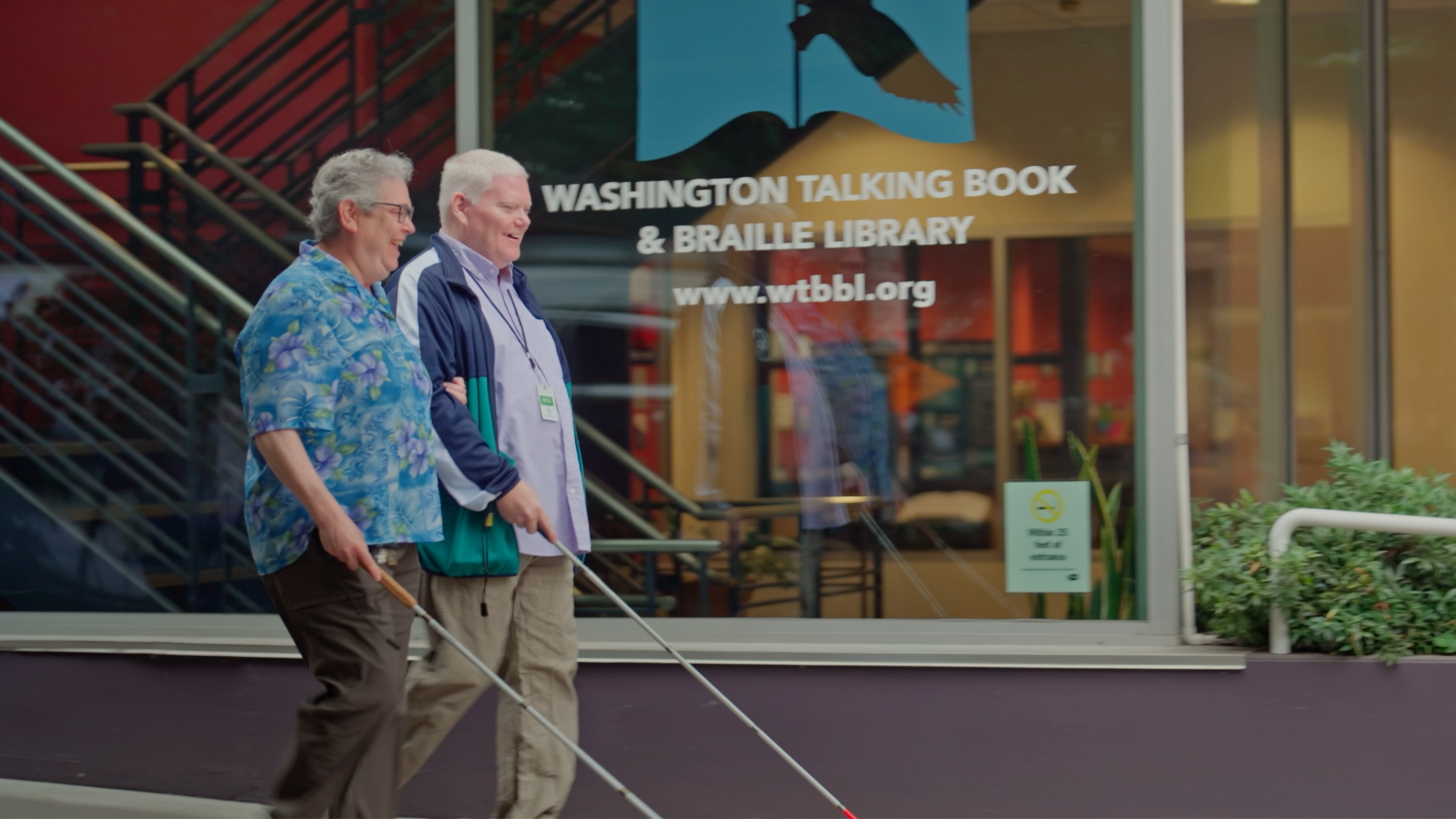 Two men using white canes are walking past the "Washington Talking Book & Braille Library" window decal